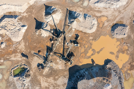 A stone crusher maschine, conveyor belts, bulldozers, dump trucks and piles of excavated stone in a quarry site seen from directly above.