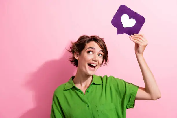 Photo of Photo portrait of lovely young lady hold heart icon look empty space dressed stylish green garment isolated on pink color background