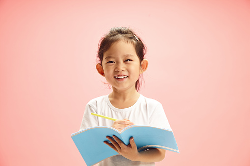 Portrait of sweet and cheerful first grader Asian ethnicity girl smiling while holding a exercise book and pen, writing something or has successfully completed the task, expresses joyful mood from getting a good mark standing over pink isolated background with a free copy space.
