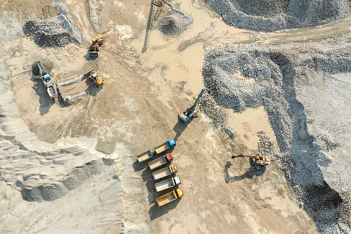 Dump trucks, bulldozers, backhoe, and heaps of excavated stone and sand in a quarry site seen from directly above.