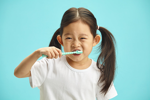 Studio shot of happy asian ethnicity cute child girl use hand holds toothbrush, brushing teeth myself at morning every day standing over blue isolated background with copy space, health medical care.