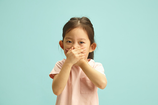 Ooops, i said it. Studio portrait of naughty korean Child Girl with Hand Over Mouth, does not want to tell the truth, tries to deceive or keep silent about something standing over blue isolated background. Keeping my secret.
