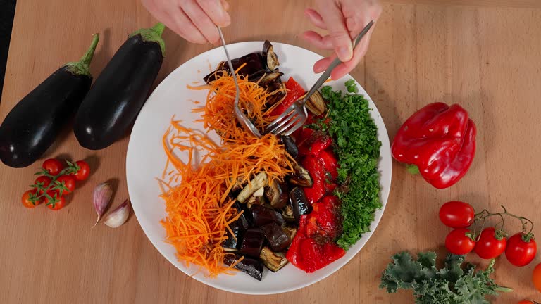 Salad of baked eggplant, paprika, fresh carrots and greens. The chef mixes vegetables and sauce on a plate, wooden table on a background of vegetables. Close-up, top view. Snack, food, vegetables, vegetable salad, eggplant dishes.