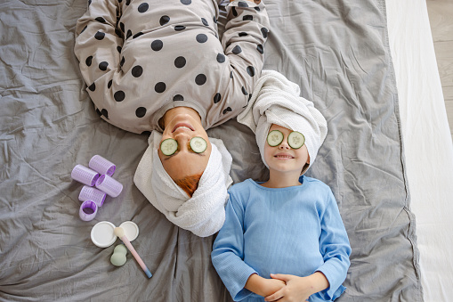 Top view of smiling mother and her little cute daughter in pajamas enjoying in bed. Their hair is wrapped in towels and they have cucumbers on their eyes