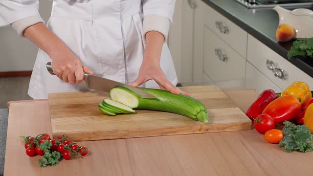 The chef cuts green zucchini into slices with a knife, a wooden table on a background of vegetables, preparing a healthy meal. Healthy eating concept, vegetarian food, vegetables. Close-up, front view.