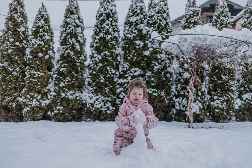 The little girl is playing in snowy weather, and trying to make a snowman. The adorable toddler in ski suit is using the snow to make a snowman outside.