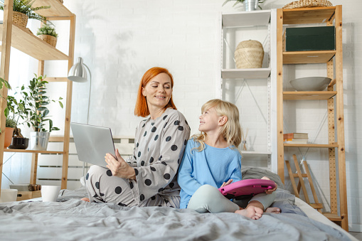 Mid adult mother using a laptop while her daughter is using a digital tablet. They are sitting on the bed and feeling cozy.