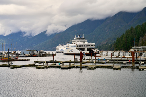 Horseshoe Bay, Canada – February 26, 2024: Two BC ferries leaving the Horseshoe Bay ferry terminal with green mountain and dramatic clouds in the background in daytime