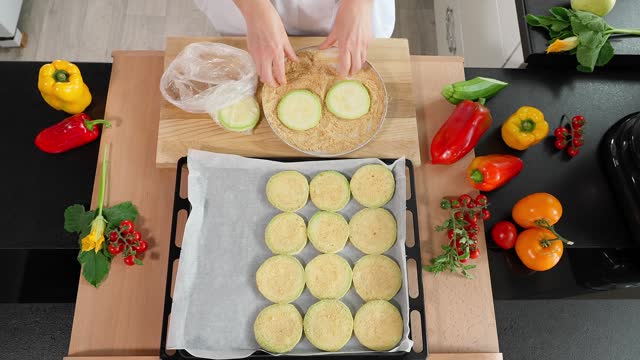 The chef sprinkles slices of white zucchini in breadcrumbs, with vegetables in the background. Zucchini dish, zucchini recipe, vegetarian food, quick snack, party. Close-up, front view, fast motion