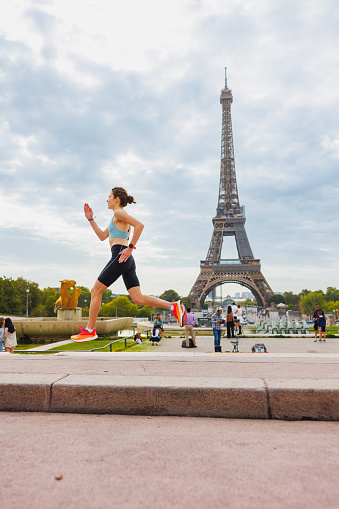 Young woman wearing sports outfit and running on a cloudy summer day in Paris, stone paved walkway near the Eiffel Tower