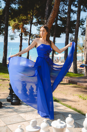 Beautiful young brunette with long brown hair wearing long blue strapless dress, standing in the park at the seaside and lifting train to the side, looking at camera, fashion and beauty