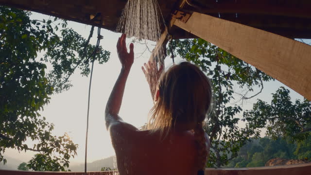 Woman takes a shower outdoors from a tree house above the jungle at sunset