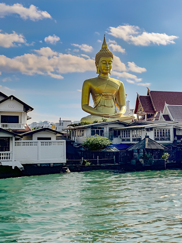 Stock photo showing  view from canal of Phra Buddha Dhammakaya Thep Mongkol, a bronze plated giant Buddha statue by Wat Paknam Bhasicharoen a royal temple, Bangkok, Thailand.