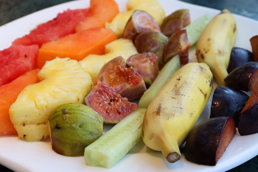 Stock photo showing a close-up, elevated view of fresh fruit salad on a white plate. The fruits pictured are slices of watermelon, papaya, pineapple, figs, melon, bananas and plum.