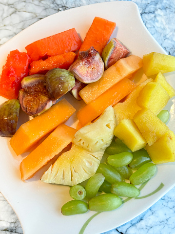 Stock photo showing a close-up, elevated view of fresh fruit salad on a white plate. The fruits pictured are slices of papaya, pineapple, figs, melon, mango and seedless white grapes.