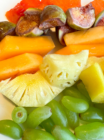 Stock photo showing a close-up, elevated view of fresh fruit salad on a white plate. The fruits pictured are slices of papaya, pineapple, figs, melon, mango and seedless white grapes.