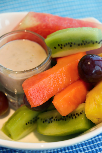 Stock photo showing a close-up, elevated view of fresh fruit salad in a white bowl surrounding glass ramekin of yoghurt. The fruits pictured are slices of papaya, pineapple, kiwi, watermelon and red grapes.