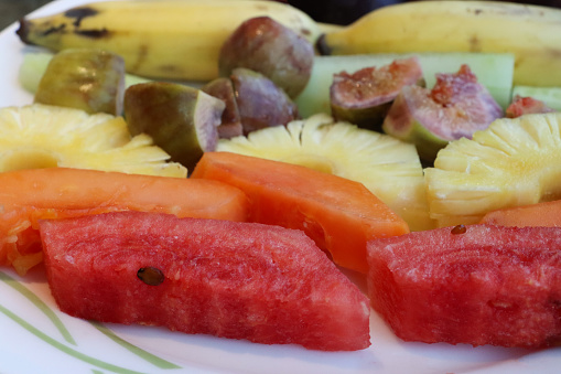 Stock photo showing a close-up, elevated view of fresh fruit salad on a white plate. The fruits pictured are slices of watermelon, papaya, pineapple, figs, melon, bananas and plum.