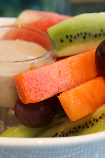 Stock photo showing a close-up view of fresh fruit salad in a white bowl surrounding glass ramekin of yoghurt. The fruits pictured are slices of papaya, pineapple, kiwi, watermelon and red grapes.