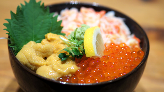 Close-up images of japanese seafood rice bowl or kaisendon sashimi donburi which have fresh ogura, uni, giant crab and topping with lemon on rice in the black bowl on the wood table from top view angle camera.