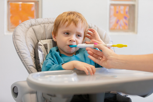 Little blond boy sitting in high chair, mother feeding him with baby spoon