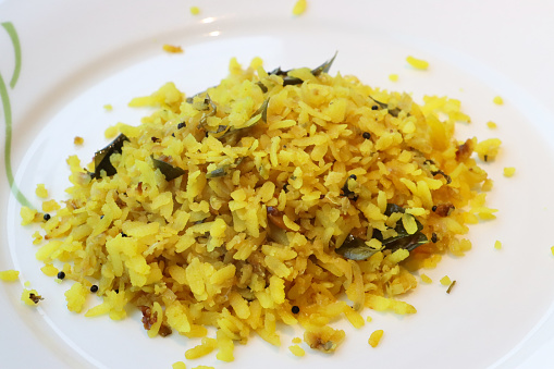 Stock photo showing close-up, elevated view of poha rice dish of spiced flattened rice with onion, green chillies, curry and coriander leaves, peanuts, mustard and cumin seeds, turmeric and lemon juice on a white plate.