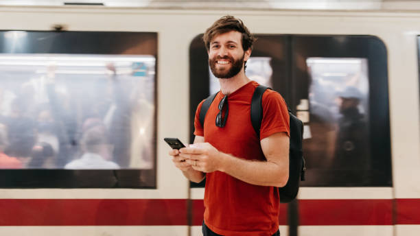 istock Bearded male commuter holding cellphone smiling cheerfully at camera beside train in underground 2117114530
