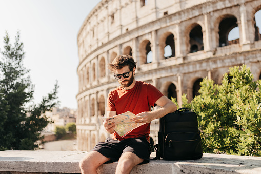 Bearded male tourist in red top and shorts, wearing sunglasses resting elbow on backpack and reading map for interesting tourist attractions, sitting on concrete wall in front of historical structure Colosseum
