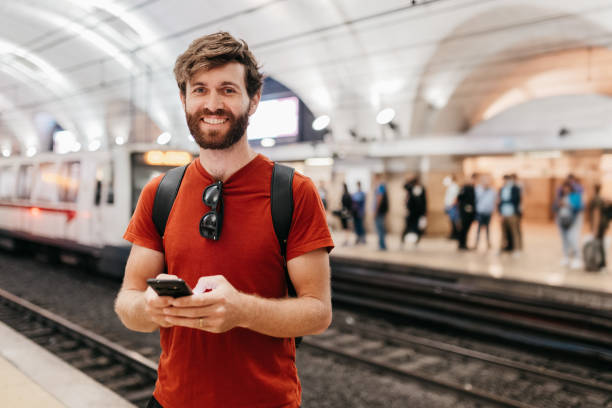 istock Bearded male commuter texting on cellphone, smiling happily at camera beside railway track in underground station 2117114541