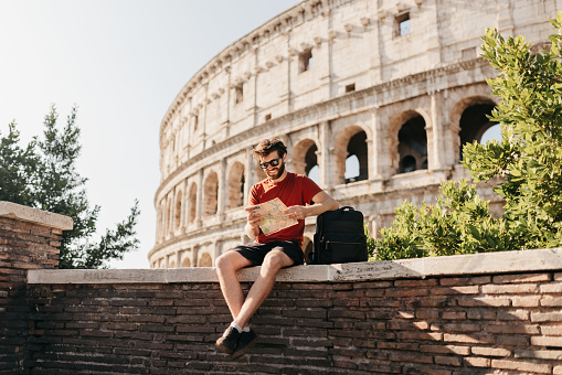 Bearded male tourist in red top and shorts, wearing sunglasses smiling on concrete wall in front of historical structure Colosseum, resting elbow on backpack and reading map for interesting tourist attractions