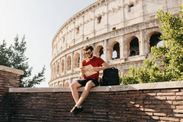 Bearded male tourist reading map and smiling with Colosseum in background Bearded male tourist in red top and shorts, wearing sunglasses smiling on concrete wall in front of historical structure Colosseum, resting elbow on backpack and reading map for interesting tourist attractions mid adult men stock pictures, royalty-free photos & images