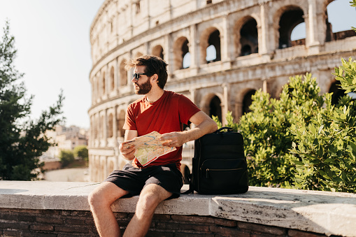 Bearded male tourist in red top and shorts, wearing sunglasses sitting on concrete wall in front of historical structure Colosseum, resting elbow on backpack and holding map, looking into distance at beautiful sceneries