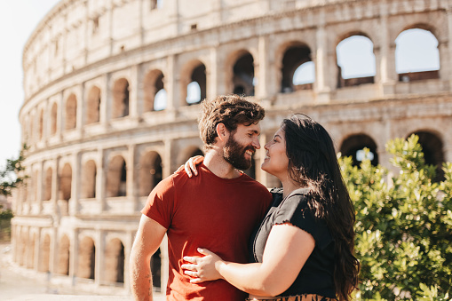 Bearded male tourist and girlfriend in casual wear hugging and smiling affectionately at each other, posing in front of historical structure Colosseum
