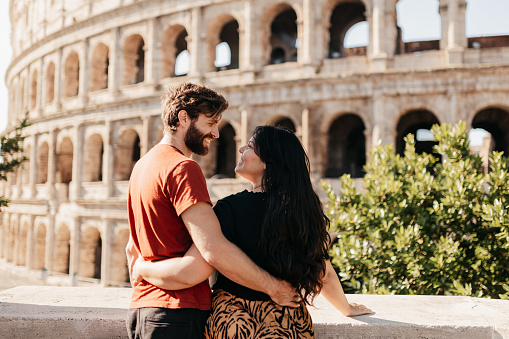 Happy bearded man and girlfriend in casual wear embracing and smiling lovingly at each other whilst on vacation, standing by stone wall in front of historical structure Colosseum