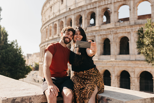 Bearded man and girlfriend in casual wear, smiling cheerfully and taking selfie with cellphone whilst on vacation, posing on stone wall in front of historical monument Colosseum