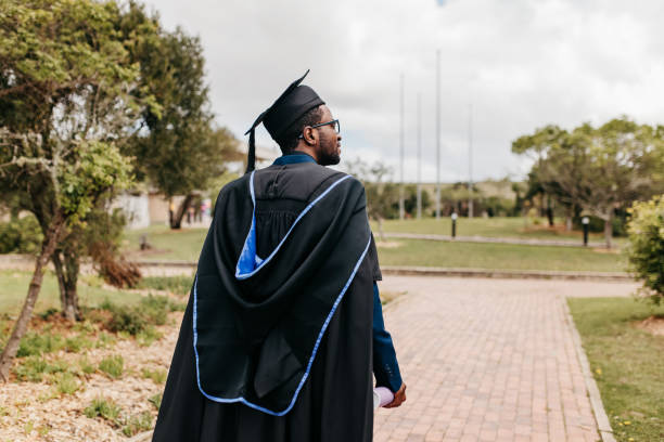 Rear view of bearded male graduate walking and looking sideways on campus grounds on graduation day Young male graduate with beard wearing graduation gown and mortarboard walking and looking sideways into distance on campus grounds graduation man stock pictures, royalty-free photos & images