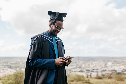 Young man with eyeglasses wearing graduation gown and mortarboard, standing on hilltop overlooking rural landscape, smiling proudly and sharing achievement on graduation day in social media with cellphone