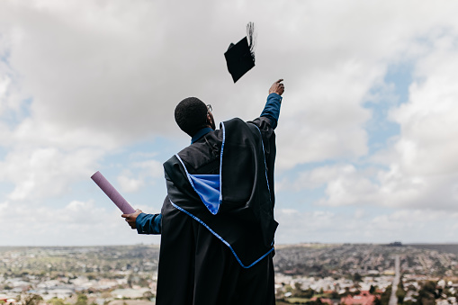 Excited man wearing graduation gown holding degree in one hand and throwing mortarboard into air on graduation day, celebrating success, standing on hilltop overlooking rural landscape on cloudy day