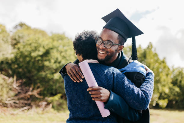 Young male graduate hugging mother on graduation day, smiling proudly at campus grounds Proud young man with eyeglasses, wearing graduation gown and mortarboard, holding degree, hugging mother and smiling happily, celebrating achievement on graduation day at campus grounds graduation man stock pictures, royalty-free photos & images
