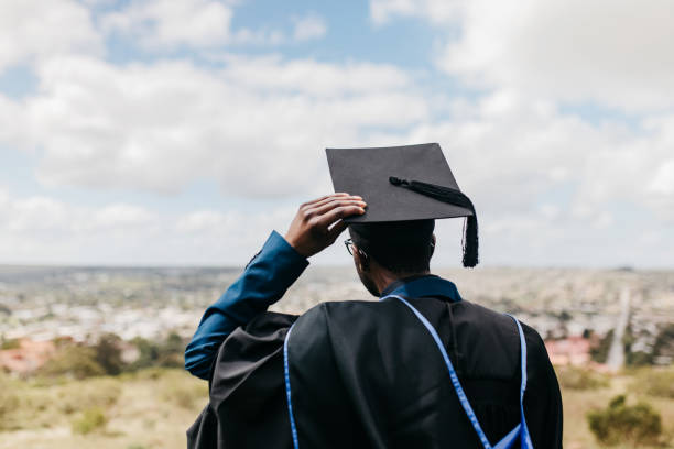 Rear view of male graduate holding onto mortarboard, looking out at rural landscape on hilltop Male graduate wearing graduation gown and holding onto mortarboard, standing on hilltop and looking out at rural landscape in distance graduation man stock pictures, royalty-free photos & images