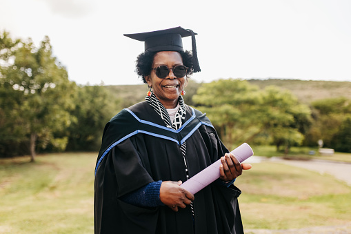 Mature woman with sunglasses, wearing graduation gown and mortarboard, holding degree, posing on graduation day and smiling proudly at camera on campus grounds