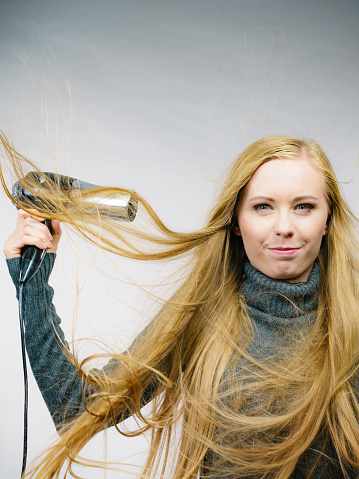Blonde woman styling her very long hair. Female with blowing messy hair holding hairdryer. Hair accessories.