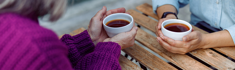 Mature women best friends with cups of fresh coffee sit at wooden table on outdoors cafe terrace on autumn day closeup