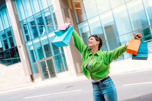 happy brunet woman in green  jacket with colorful bags having fun time with shopping in urban city. Consumerism, sale, purchases, shopping, lifestyle concept