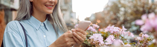 istock Smiling grey haired Asian woman uses cellphone standing on outdoors terrace 2117140231