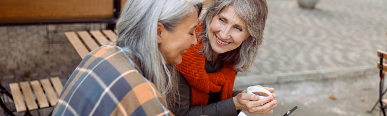 Long haired mature Asian woman shows mobile phone to best friend with cup of coffee at small table in street cafe on autumn day