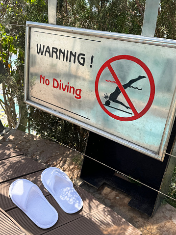 Stock photo showing close-up view of swimming pool paved side with no diving warning sign for health and safety to prevent accidents and potential drowning hazard for swimmers.