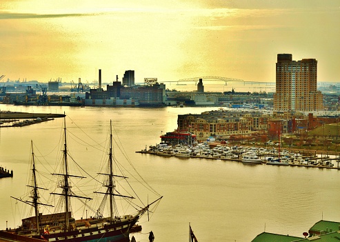 A golden sunrise on Baltimore's Inner Harbor. A scene filled with icons including The USS Constellation Sloop of War (circa 1854), the Domino Sugar building who's neon sign lights up the harbor skyline, the Harborview building and off in the distance, the Francis Scott Key Bridge, marking the area where Francis Scott Key penned the poem which became the basis for our National Anthem, The Star Spangled Banner.