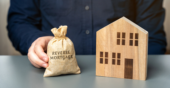 Reverse mortgage concept. Allowing homeowners to borrow money by using their home as collateral for the loan. Money bag in the hands of a businessman and a wooden house