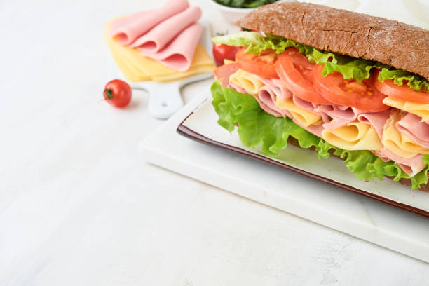 istock Sandwich. One fresh big submarine sandwich with ham, cheese, lettuce, tomatoes and microgreens on light background. Healthy breakfast theme concept, school lunch, breakfast or snack. 2117185930